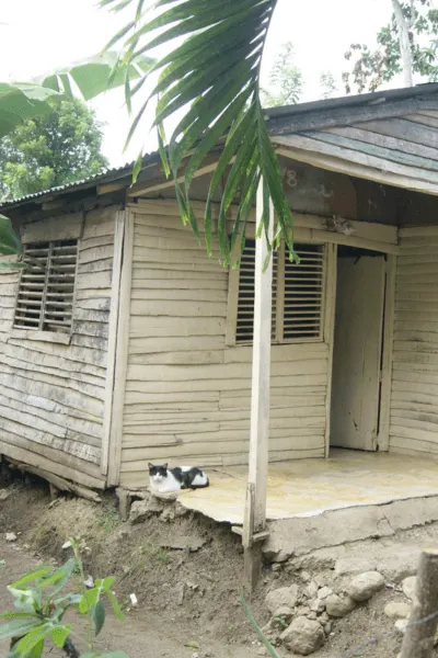 Casa antigua de madera en estado de deterioro, con un gato en la entrada. Representa el estado original de la vivienda antes de ser reconstruida.